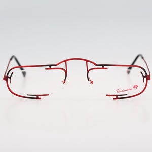 May include: Red and black rimless eyeglasses with a thin metal frame and clear lenses. The brand name "Cazal" is printed on the right lens.