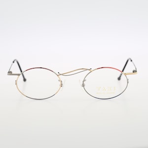 May include: Oval eyeglasses with a gold-coloured frame and black temples. The top of the frame has a red accent. The lenses are clear, and the text "TAXI" is visible on the right lens. The glasses are set against a white background.