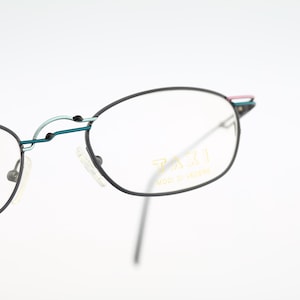 May include: Black oval-shaped eyeglasses with a thin metal frame. The glasses have a teal bridge and accents of pink and teal on the temples. The text "TAXI MOD. & DESIGN" is printed on the lens.