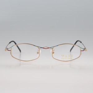 May include: Gold-tone rimless eyeglasses with a blue and red accent on the frame. The brand name "TAXI" is printed on the right temple.