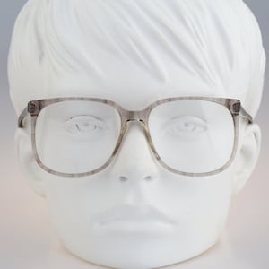 May include: Gray square-framed eyeglasses with clear lenses. The frames have a marbled appearance and are set against a white head form. The glasses have a classic, retro style.