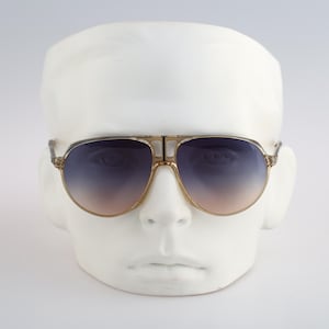 May include: A pair of gold-rimmed aviator sunglasses with gradient blue and brown lenses. The sunglasses are on a white mannequin head.
