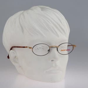 May include: Oval-shaped eyeglasses with thin gold-tone metal frames and tortoiseshell-patterned temples. The right lens features the text "CONTROVERSE" printed in a reddish-orange font. These glasses have a classic bridge and nose pads.