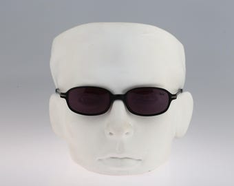 May include: Black oval-shaped sunglasses with dark tinted lenses and thin black frames. The glasses feature a small white logo on the upper corner of the right lens. The frames have a simple, streamlined design with narrow temples extending from the sides.