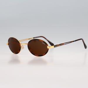 May include: Oval sunglasses featuring brown lenses and a gold-tone metal frame. The temples are thin with a tortoiseshell pattern and gold accents. The brand name "Krizia" is printed on the right lens and on the side of the temple arms.