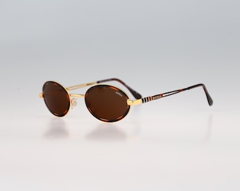 May include: Oval sunglasses featuring brown lenses and a gold-tone metal frame. The temples are thin with a tortoiseshell pattern and gold accents. The brand name "Krizia" is printed on the right lens and on the side of the temple arms.