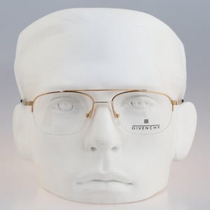 Puede incluir: Gafas de metal en tono dorado con diseño de doble puente y montura semi-al aire. El cristal derecho presenta el texto impreso en negro GIVENCHY con un pequeño logotipo encima. Las gafas se muestran sobre una cabeza de maniquí blanca.