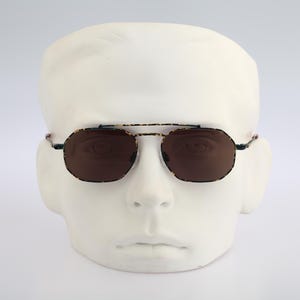 Puede incluir: Gafas de sol estilo aviador con doble puente y lentes marrones. Las monturas tienen un patrón de carey. Las gafas de sol se exhiben en una cabeza de maniquí blanca, mostrando el diseño y el ajuste.