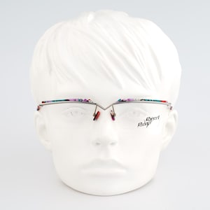 May include: Silver-tone semi-rimless spectacles featuring a V-shaped bridge and multicoloured patterned accents along the top brow bar. The right lens displays the text Robert Rudger in black script. The nose pads are a distinct red colour.