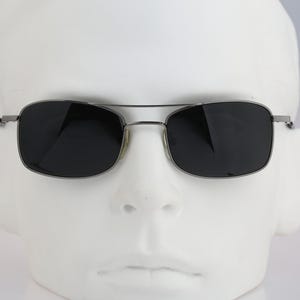 May include: Rectangular sunglasses featuring a thin, dark metallic frame and a double bridge design. The lenses are dark, solid black, providing full coverage. The frame has slim, straight temples that extend from the outer corners of the lenses.