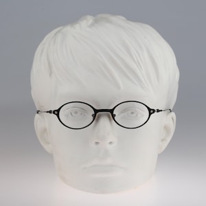 May include: A pair of black oval-shaped eyeglasses with thin metal frames and a bridge, displayed on a white mannequin head. The glasses feature slender temples that extend from the sides of the oval rims, providing a clear view of the facial features.