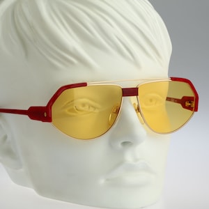May include: Retro-style sunglasses with a gold metal frame, red accents, and yellow lenses. The glasses have a unique geometric shape and are displayed on a white mannequin head. A stylish accessory.