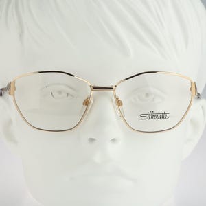 Könnte beinhalten: Goldfarbene Metallbrille mit einem dünnen, kantigen Rahmendesign. Das rechte Brillenglas trägt den Markennamen Silhouette in schwarzer Schreibschrift. Die Brille wird auf einem weißen Schaufensterpuppenkopf präsentiert.