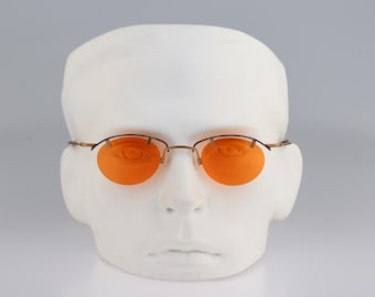 May include: Oval-shaped glasses featuring vibrant orange lenses and a thin, metallic gold-toned frame. The design includes a distinctive brow bar that connects the upper edges of the lenses, supported by slender temples and a central bridge.