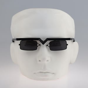 May include: Black sunglasses featuring a distinct V-shaped bridge design and rectangular lenses. The frame has a straight top bar that connects to the temples, while the dark lenses are suspended below the frame, creating a unique layered appearance.