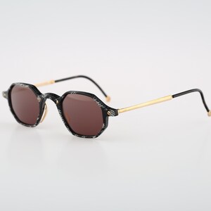Puede incluir: Gafas de sol octogonales con montura estampada en blanco y negro y lentes marrones. Las patillas son doradas con puntas negras. Un accesorio elegante para la protección ocular.