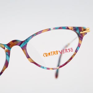 May include: Cat-eye glasses with a multicolored tortoiseshell pattern in shades of purple, teal, and gold. The clear lenses feature the orange text "CONTROVERSE" printed on the right side. The frame includes gold-toned metal hinges.