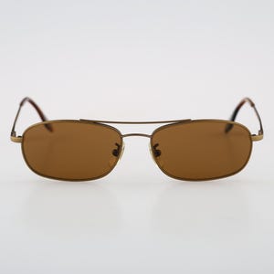 May include: A pair of sunglasses with a thin, matte gold-toned metal frame and a double bridge design. The lenses are a solid, medium-brown tint, and the temples feature thin metal arms with dark, tortoise-shell patterned tips at the ends.