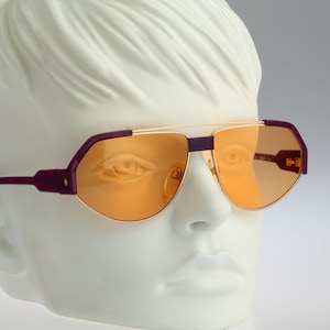 May include: Purple and gold aviator sunglasses with orange lenses. The sunglasses have a unique, angular shape.