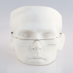 Puede incluir: Gafas sin montura de color plateado con una estructura de metal fina y lentes rectangulares. La lente derecha muestra el texto "KABANK" en letras negras y naranjas. Las gafas se presentan sobre una cabeza de maniquí blanca, destacando el diseño minimalista y la construcción ligera de la montura.