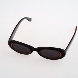 May include: Black oval sunglasses with brown tinted lenses. The sunglasses have a thin black frame.
