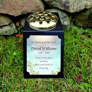 May include: Black granite memorial with a gold flower vase and a white cross. The inscription reads "In Memory of Our Dad David Williams 1950-2023 Those we love don't go away. They walk beside us every day."