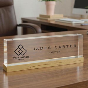 May include: A clear acrylic desk nameplate with a wood grain design. The nameplate features the name "JAMES CARTER" and the title "LAWYER" in black text. A custom logo is displayed on the left side. The base is gold.