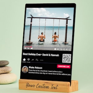 May include: A black digital photo frame with a wooden base displays a video titled "Best Holiday Ever - David & Hannah." The video shows a couple on swings over the ocean. The wooden base has the text "Your Custom Text."