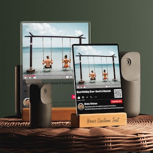 May include: Two tablet devices displaying a video of a couple on a swing set over a body of water. The video title is "Best Holiday Ever - David & Hannah". The tablets are on wooden stands with a speaker in between them. The text "Your Custom Text" is on the wooden stand in front of the speaker.