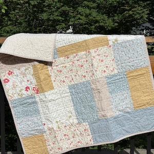 May include: A patchwork quilt featuring rectangular fabric panels in various patterns and colors, including mustard yellow, light blue, and white with red floral prints. The quilt has visible grid-like stitching throughout the entire surface.