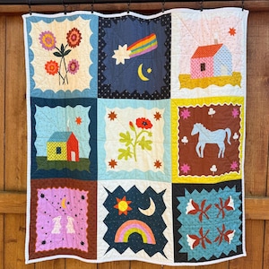 May include: A handmade quilt with nine colorful squares, each featuring a unique design. The squares depict flowers, a rainbow, a house, a horse, and other whimsical elements. The quilt is bordered with a white scalloped edge and hangs against a wooden fence.