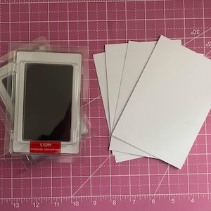 May include: A baby hand and footprint kit with a black ink pad and several white cards. The ink pad is in a clear plastic package with a red "STOP!" label. The items are on a pink cutting mat.