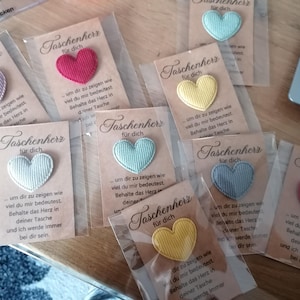 Könnte beinhalten: Neun kleine Karten mit dem Text "Taschenherz für dich" und einem kleinen gestickten Herz in verschiedenen Farben, darunter Rot, Gelb, Blau und Grün. Die Karten sind in Klarsichtfolie verpackt.