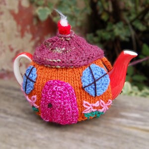 May include: A ceramic teapot covered in a hand-knitted orange cozy designed to look like a small house. The cozy features a dark pink roof, a bright pink crocheted door, two light blue circular windows, and small pink and green floral details.