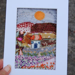 May include: A textile art piece featuring a landscape scene with a small house, a bright orange sun, and various floral patterns. The artwork is created with layered fabric patches and intricate embroidery stitches, all framed within a white mat.