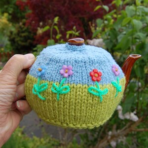 May include: A hand-knitted tea cosy in blue and green, decorated with flower appliqués. The cosy has a light blue top and a green bottom, with a brown spout and a brown knob. The flowers are pink, red, and blue. A charming, handmade item.