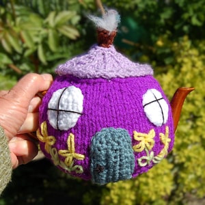 May include: A hand-knitted teapot cosy in purple, lavender, and green, resembling a house. It features white window details, a teal door, and yellow floral embroidery. The design is completed with a brown spout and chimney, perfect for a tea party.
