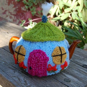 May include: A light blue knitted tea cosy shaped like a small house with a green roof, a bright pink door, two yellow circular windows with black cross stitching, and red flower accents at the base, topped with a small blue knob and white tuft.