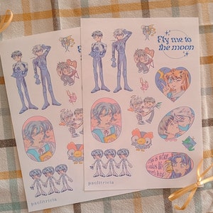 May include: Two sheets of stickers featuring anime characters. The stickers are on a light pink background with a white border. The first sheet features a character with blue hair and a blue and white outfit. The second sheet features a character with pink hair and a pink and white outfit. The text "Fly me to the moon" is on the second sheet.