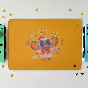Pode incluir: Um tapete de rato amarelo com uma ilustração de desenho animado de um animal preto e branco usando um laço vermelho e um boné. O animal está rodeado de estrelas e flores. O tapete de rato está ao lado de dois controladores Nintendo Switch, um verde e um azul.