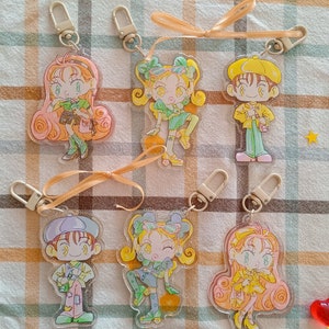 Könnte beinhalten: Sechs bunte Schlüsselanhänger mit verschiedenen Designs von Comicfiguren. Jeder Schlüsselanhänger hat einen Metallring und eine transparente Plastikrückseite. Die Figuren tragen alle verschiedene Outfits und haben verschiedene Frisuren.