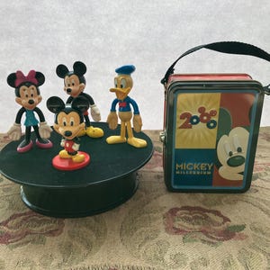 May include: A set of four Disney character figurines, including Mickey Mouse, Minnie Mouse, Donald Duck, and Goofy. The figurines are standing on a black turntable. A red and green tin lunchbox with the text "Mickey Millenium 2000" is in the background.