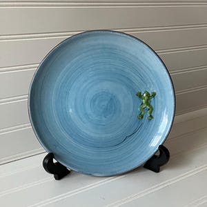 May include: A round, light blue ceramic plate with a dark brown rim. The plate features a green frog figurine and a black line. The plate is displayed on a black stand against a white background.