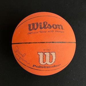 May include: An orange Wilson basketball with black text that reads "Wilson Official Size and Weight." The basketball has a white "W" logo and a signature. The basketball is on a black surface.