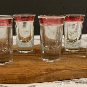 May include: Six clear glass shot glasses with a red and white band around the top. The glasses are sitting on a wooden cutting board.