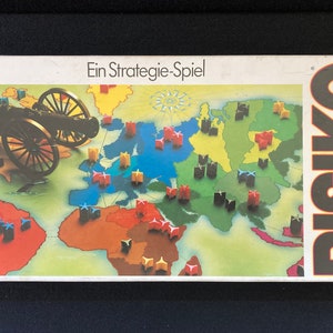 May include: A vintage board game box with a world map and a cannon on the front. The box is white with black text that reads "Ein Strategie-Spiel" and "RISIKO".
