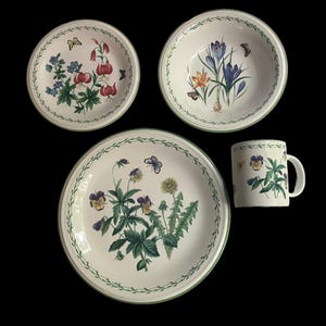 May include: A set of white ceramic tableware featuring floral and butterfly designs. The set includes a bowl, a plate, and a mug. The rims are decorated with a green vine pattern. The floral designs include red, blue, and purple flowers.