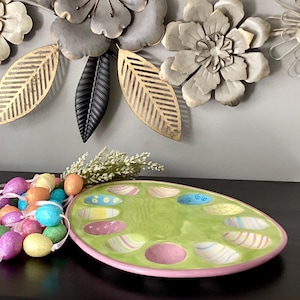 May include: An oval-shaped, green ceramic Easter egg serving platter with a pink rim. The platter has egg-shaped indentations with pastel designs. A garland of colorful glitter eggs and decorative flowers are nearby.