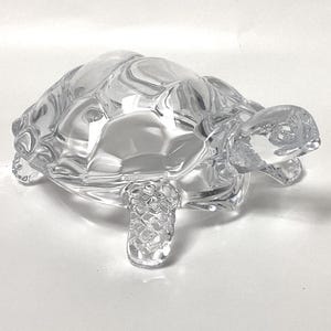 May include: A clear glass turtle figurine with detailed shell and limbs. The figurine is transparent, allowing light to pass through, and sits on a white surface. The turtle's design includes textured patterns on its shell and legs.
