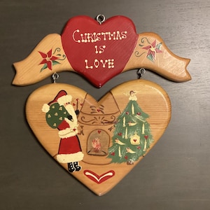 May include: Wooden Christmas heart ornaments. The top heart is red with the words "Christmas is Love." The bottom heart features Santa Claus, a fireplace, and a Christmas tree. The ornaments are painted with festive colors.